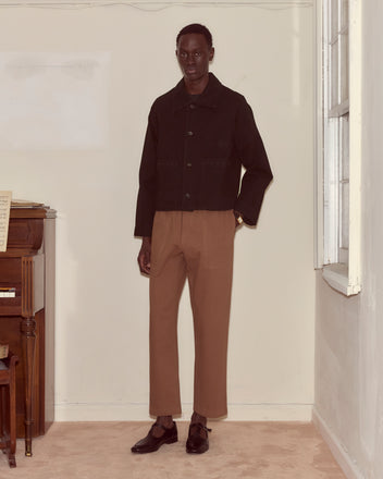 A person stands indoors next to a piano, wearing an Embroidered Denim Jacket - Black with a boxy fit, brown pants, and dark shoes.