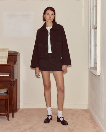 A woman in an embroidered black denim jacket stands indoors by a piano, paired with a skirt, white socks, and black shoes.