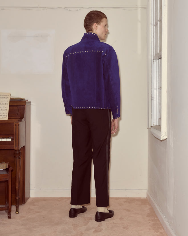 Person stands indoors facing away, wearing the Studded Welding Jacket - Blue, paired with black pants and black-and-white shoes.