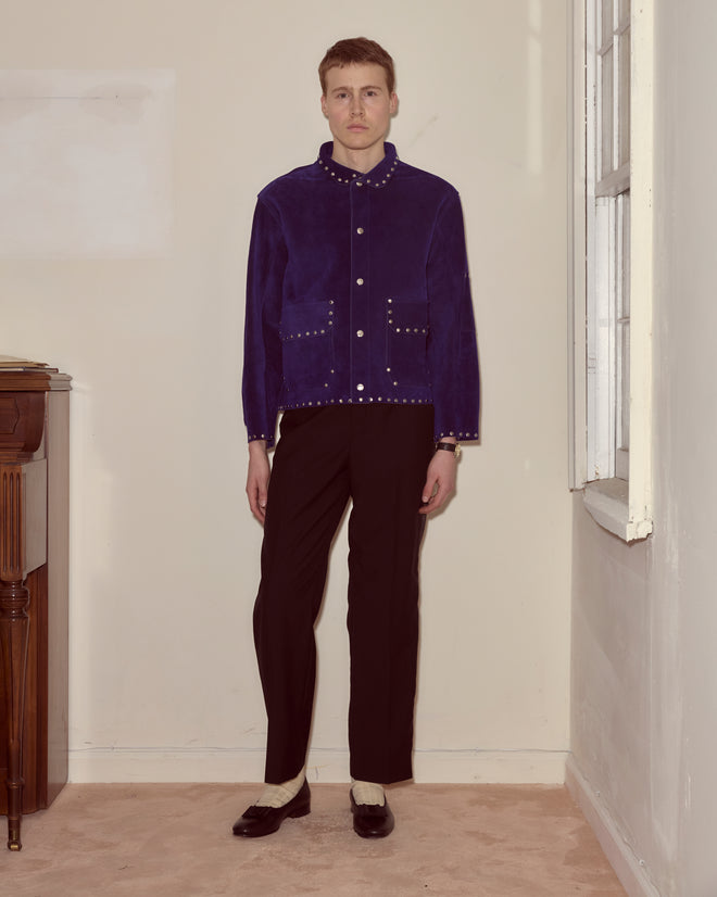 A person wearing a Studded Welding Jacket - Blue and black pants stands in a plain room next to a window.