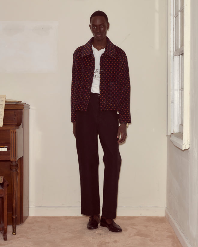 Indoors by a piano, a person wears the McAllister Corduroy Jacket with a foulard pattern, paired with a white shirt and black pants.