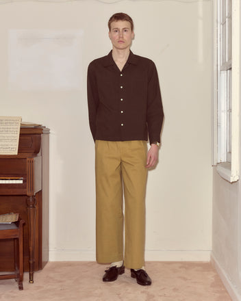 A person in a brown Voile Long Sleeve Shirt and tan pants stands indoors beside a piano, looking directly at the camera.