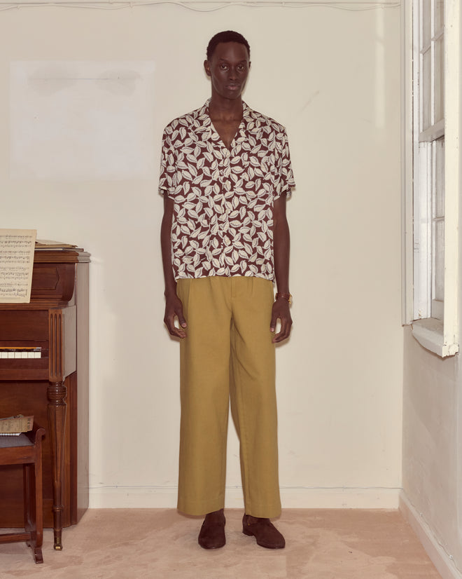 A man stands indoors by a piano, wearing the Paper Birch Short Sleeve Shirt with a leaf-stamp pattern and mustard wide-leg trousers.