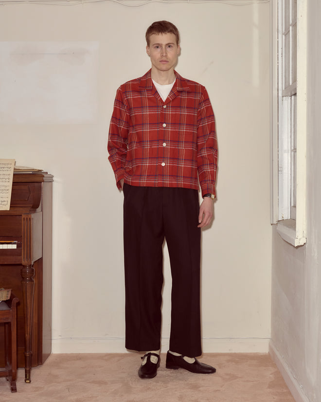 A man wearing the Gable Plaid Overshirt and black trousers stands indoors beside a piano and a window.
