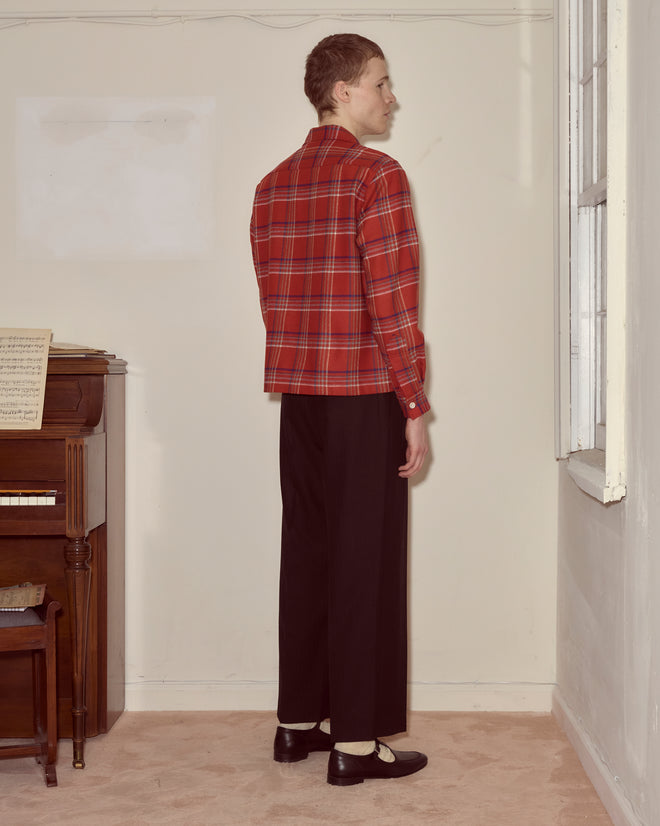 A person wearing a Gable Plaid Overshirt and black pants stands facing a corner near a piano and a window.