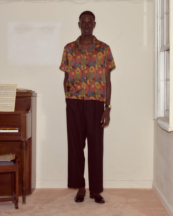 A person wearing the Fruit Pit Short Sleeve Shirt and black pants stands indoors beside a piano and window.