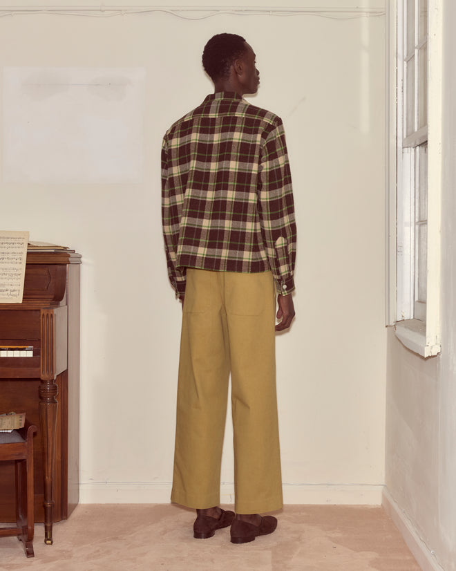 Indoors near a piano, a person faces a wall wearing the Woodcrest Overshirt in brown and green plaid with wide-leg beige pants.