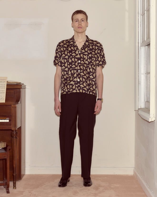 A person stands indoors by a piano, facing the camera and wearing the Paper Hat Shirt with black trousers.