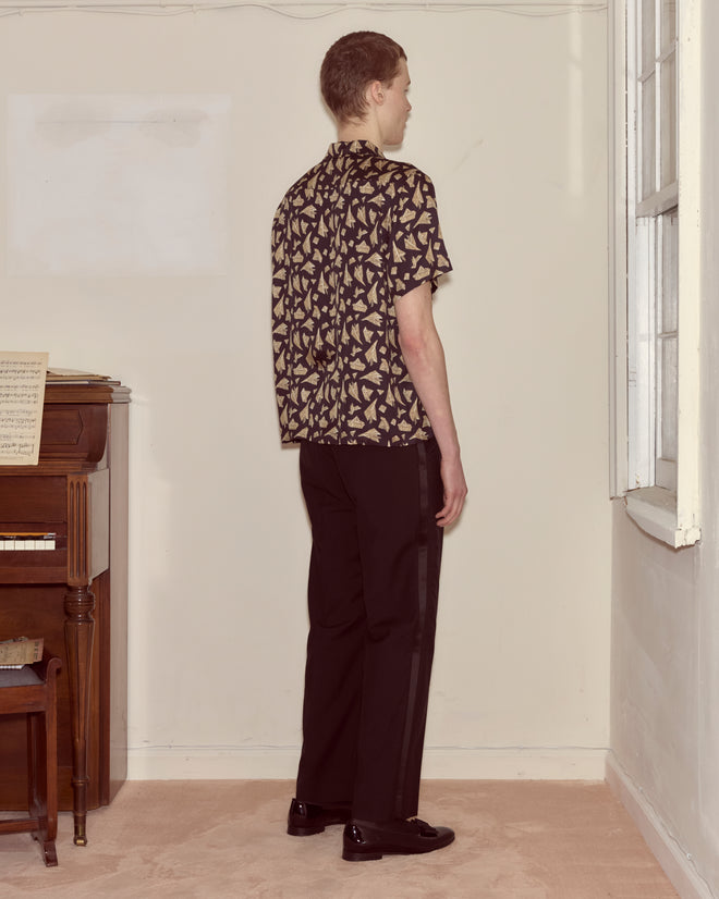 Wearing the Paper Hat Shirt and black pants, a person stands by a piano facing a window in a simple room.