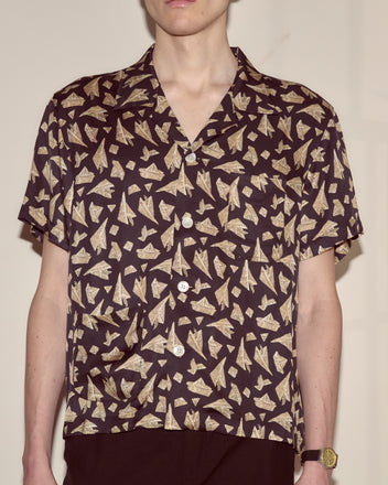 A person wearing the Paper Hat Shirt, a short-sleeved top featuring a novelty beige paper airplane pattern, with a wristwatch, stands against a plain wall.