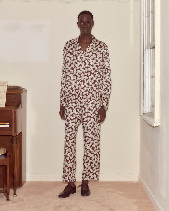 A person stands indoors beside a piano and window, wearing the Paper Birch Long Sleeve Shirt—viscose pajamas with a leaf-stamp pattern and Peter Pan collar—paired with dark shoes.