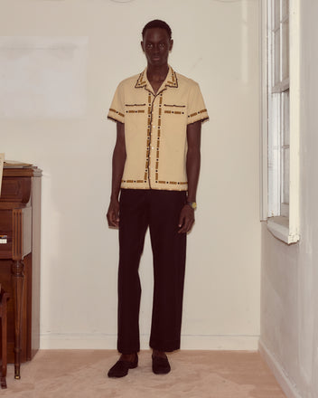 A man stands indoors wearing the Beaded Ryegrass Short Sleeve Shirt, featuring handmade beadwork, paired with black pants beside a piano and window.