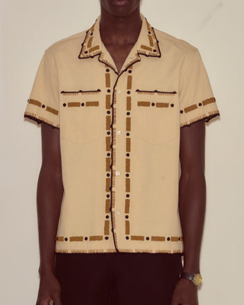 A person models the Beaded Ryegrass Short Sleeve Shirt, featuring beige fabric and brown and black stitched patterns, while standing in front of a plain wall.