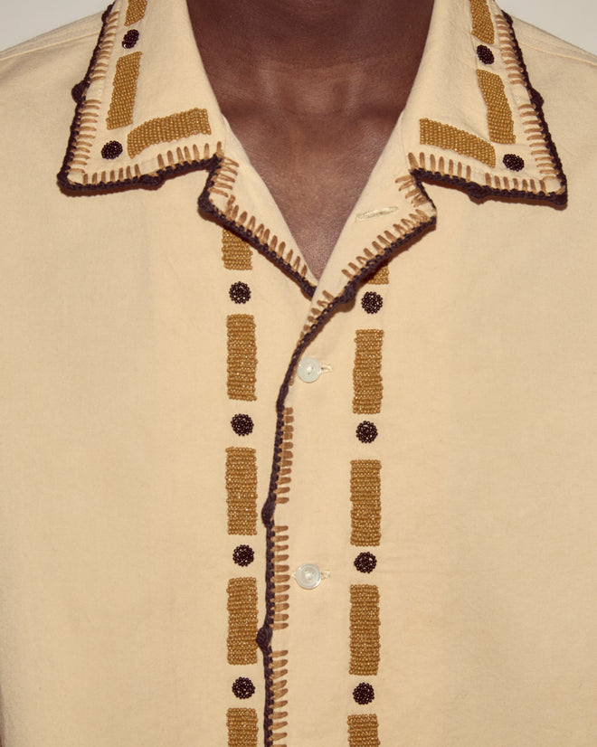 The Beaded Ryegrass Long Sleeve Shirt features handmade cotton fabric with brown and yellow embroidery and bronze bead accents, shown on a model with medium brown skin.