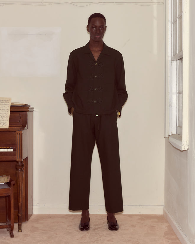 Wearing the Astor Trillium Shirt in Black and a dark suit, a person stands indoors by a piano with hands in pockets, facing the camera.