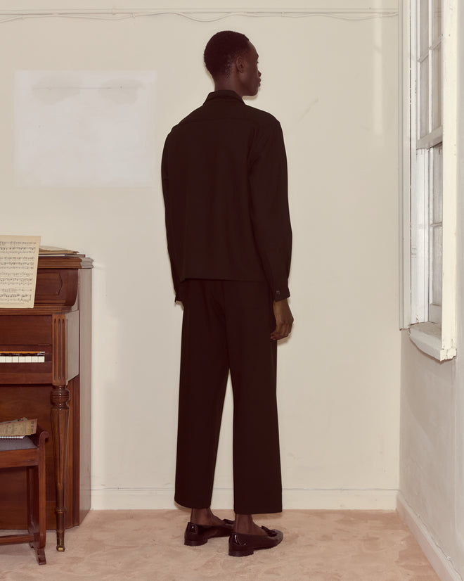 A person in black, wearing the Astor Trillium Shirt - Black, stands facing a wall near a window and an upright piano with sheet music.