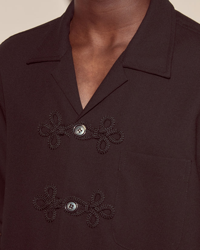 A close-up of a person wearing the Astor Trillium Shirt in Black, featuring decorative frog closures and a chest pocket.