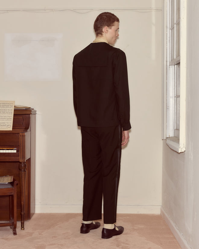 Wearing the Shadow Cricket Shirt, a person stands facing a wall near a piano and window in a softly lit room.