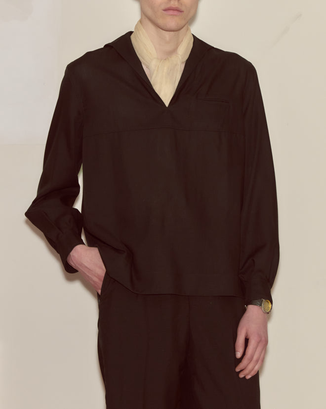A person wearing the Shadow Cricket Shirt—a loose black shirt with a sailor collar—along with black pants, a cream scarf, a watch, and one hand in their pocket.