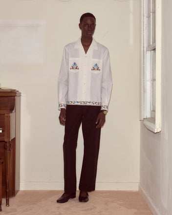 A man wearing the Nouveau Vine Long Sleeve embroidered white shirt with blue patterns and black pants stands by a window in a softly lit room.