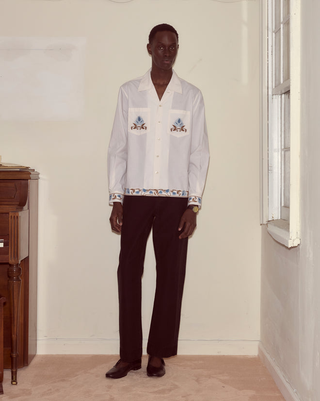A man stands indoors by a window and wooden piano, wearing the Nouveau Vine Long Sleeve, a white embroidered floral shirt made from 100% cotton, paired with black pants and black shoes.