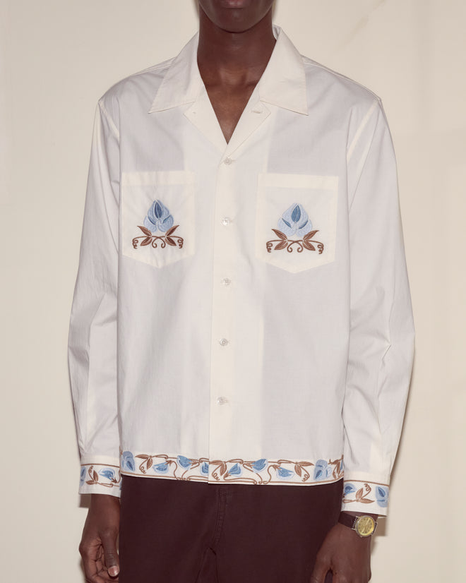 A man wears the Nouveau Vine Long Sleeve, a white 100% cotton poplin shirt with blue floral embroidery on the pockets, hem, and cuffs.