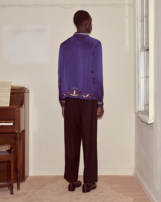 Dressed in the Dream Souvenir Long Sleeve and black pants, a person stands facing a wall near a piano and window in a softly lit room.