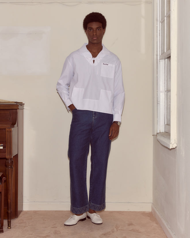 A man in a Cotton Maze Long Sleeve Shirt and blue jeans stands in a neutral-colored room beside a piano and window.
