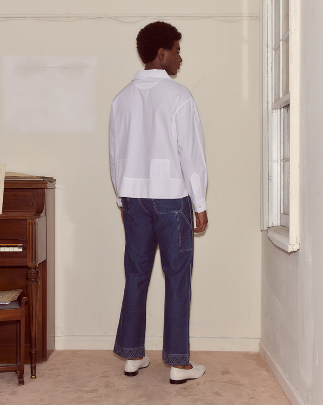 A person in a Cotton Maze Long Sleeve Shirt and blue jeans stands indoors facing a beige wall near a window and piano.
