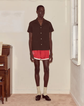 A man in a brown Voile Short Sleeve Shirt with a boxy silhouette and red athletic shorts stands indoors near a piano and a window.