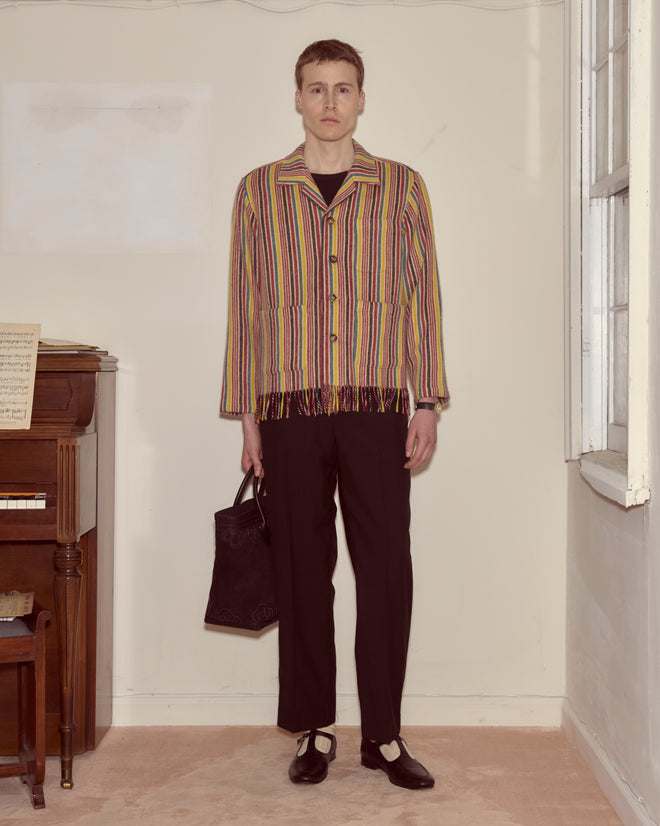 A person stands in a room by a piano, wearing the Lawrence Overshirt with a stripe pattern and black pants, holding a black bag.