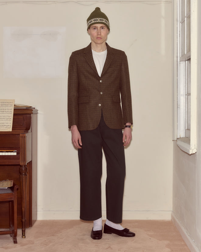 Wearing the Morris Houndstooth Suit Jacket, a person stands indoors by a piano and window, styled with a beanie, t-shirt, and loafers.