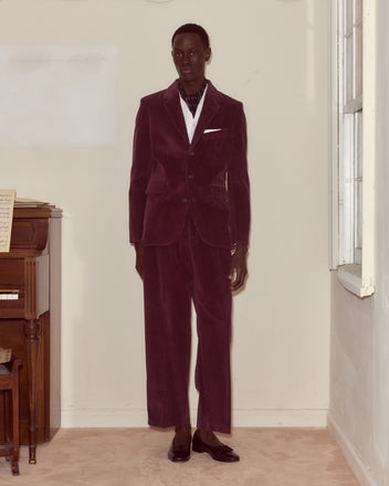 A man in a dark purple Corduroy Suit Jacket stands indoors by a piano and window, paired with black shoes and a white shirt, capturing the refined style of 1950s men's fashion.