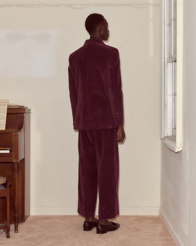 A person in a Corduroy Suit Jacket - Dark Purple stands by a piano and window, facing the corner of a room and showcasing classic 1950s men's style.