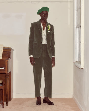 A person in a green Corduroy Suit Jacket stands indoors by a piano near a sunlit window, channeling the refined elegance of 1950s suiting.