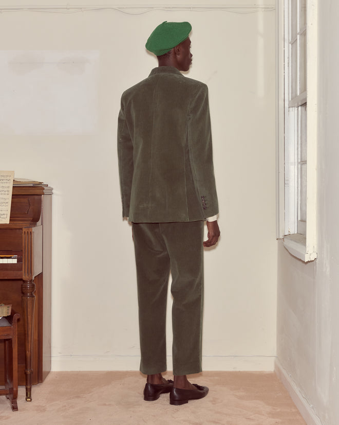 A person in a green Corduroy Suit Jacket stands facing a wall beside a piano in a sparsely furnished room.