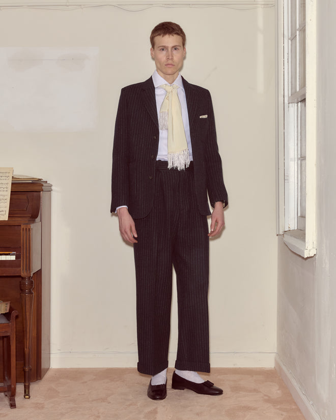 A man in the Waldorf Pinstripe Suit Jacket stands indoors by a window and piano, pairing classic style with a fringe scarf.