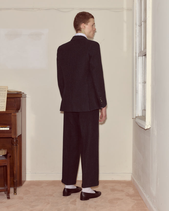 A person in a Waldorf Pinstripe Suit Jacket stands indoors facing a window, with a piano and sheet music visible in the background.