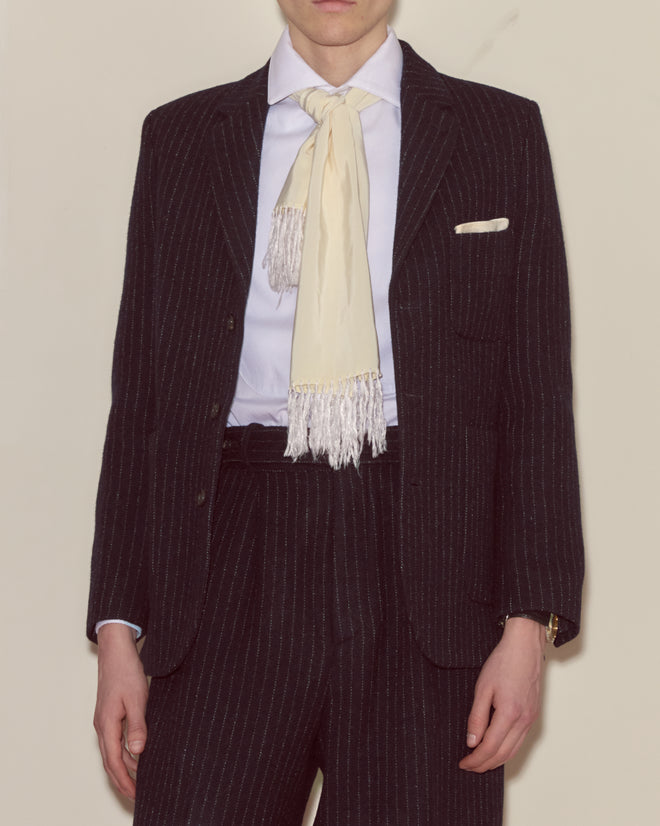 Person in a heavyweight wool suit featuring the Waldorf Pinstripe Suit Jacket, paired with a white shirt and cream fringed scarf, standing against a plain background.
