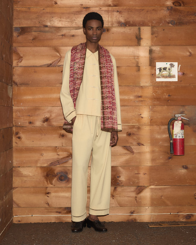 Man in cream outfit and Arrowleaf Scarf stands by wooden wall with fire extinguisher and small dog photo.