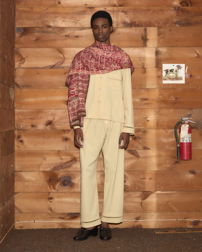 A person in a cream-colored outfit with the patterned red Arrowleaf Scarf, standing by a wood wall, fire extinguisher, and dog photo.