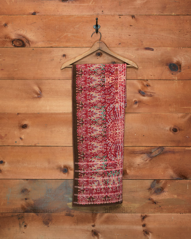 The Arrowleaf Scarf, crafted from soft wool and adorned with a red paisley pattern, hangs on a wooden hanger against a wooden wall, evoking the intricate designs of traditional Kashmiri shawls.