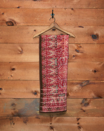The Arrowleaf Scarf, crafted from soft wool and adorned with a red paisley pattern, hangs on a wooden hanger against a wooden wall, evoking the intricate designs of traditional Kashmiri shawls.