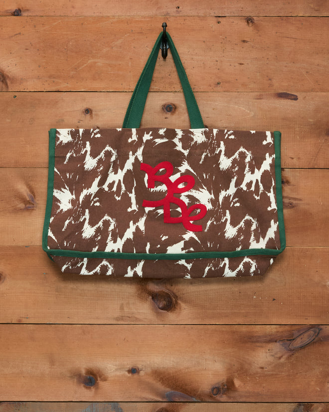The Pony Graphic Tote, an oversized cotton bag featuring a brown and white pattern, green straps, and bold red “Be Be” text reminiscent of an Appaloosa pony, hangs on a wooden wall.