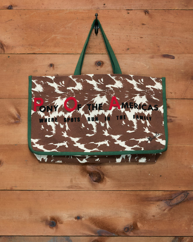 Pony Graphic Tote—an oversized cotton bag in a brown and white pattern with “PONY OF THE AMERICAS” text, displayed hanging on a wooden wall.