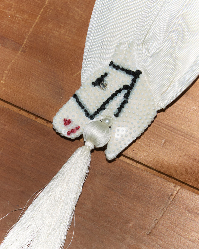 Sequin Pony Scarf - Cream displayed on a wooden surface, featuring a beaded white silken tassel and sequined horsehead design.