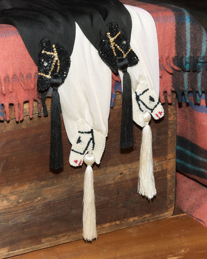 Three beaded horse-head bolo ties with tassels hang over a Sequin Pony Scarf - Black, draped across a wooden chest.