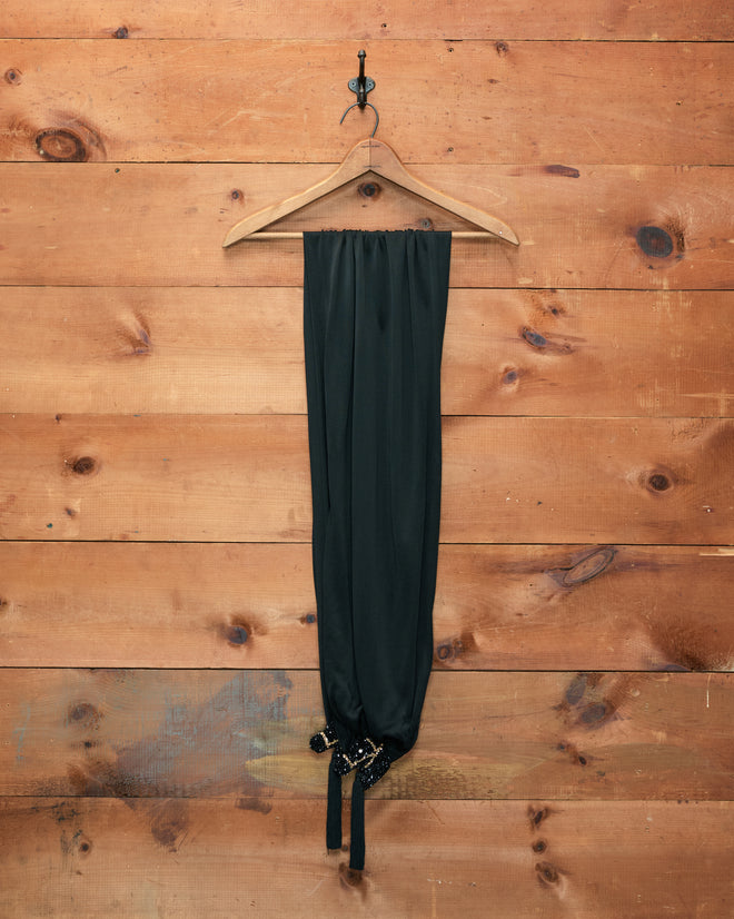 Black pants on a wooden hanger hanging from a hook against a wooden plank wall background, complemented by the Sequin Pony Scarf - Black, draped elegantly nearby.