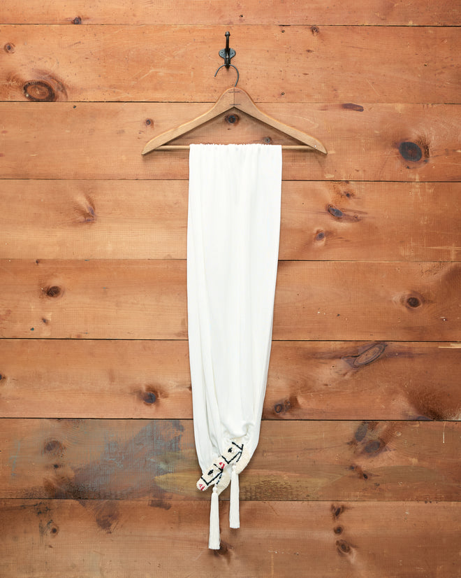 The Sequin Pony Scarf in Cream hangs on a wooden hanger against a wooden wall, tied at the bottom with two knots, its simple elegance reminiscent of a silken tassel.