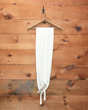 The Sequin Pony Scarf in Cream hangs on a wooden hanger against a wooden wall, tied at the bottom with two knots, its simple elegance reminiscent of a silken tassel.